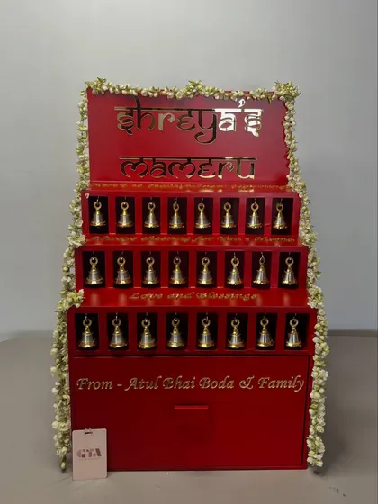 This custom-built structure was designed for a 'Mameru' ceremony, a cherished tradition of gifting from the maternal family to the bride. The vibrant red is accented with gold lettering in Hindi and delicate jasmine garlands, with each niche holding a small, symbolic bell.