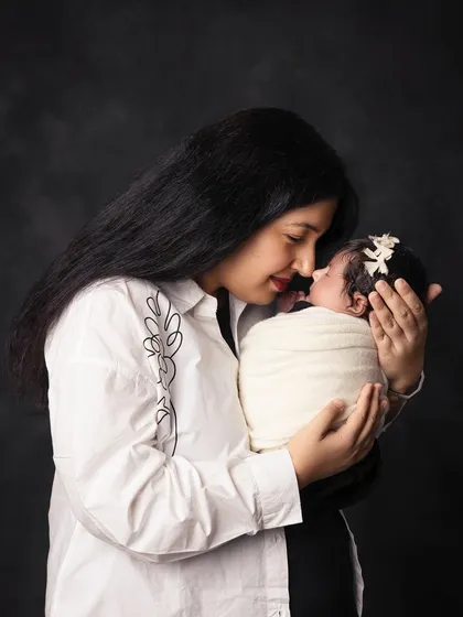 A close-up portrait of a mother and her newborn against a dark, classic background. The focus is entirely on her loving expression and the baby's peaceful sleep.