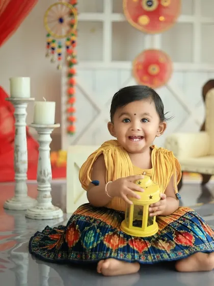 A baby girl's bright smile lights up this Diwali themed photo. She is sitting on the floor in a colorful traditional dress, holding a yellow lantern, with a beautifully decorated studio background.