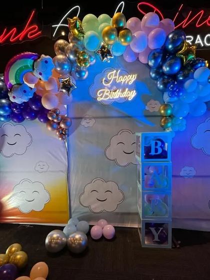 A dreamy sky theme for a first birthday with smiling cloud backdrops. The setup is complete with a multi-colored balloon arch, a 'Happy Birthday' neon sign, and a light-up number '1'.