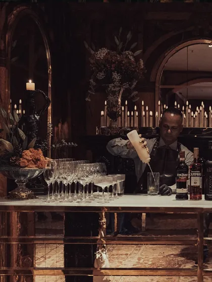 A bartender crafting cocktails at a bespoke bar, set against the moody, candlelit backdrop of our Versailles-themed event. This captures the element of professional, seamless service that complements our elaborate event designs.