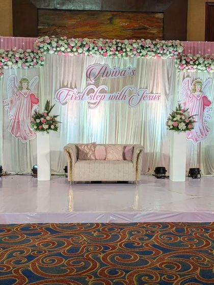 The main stage for the First Holy Communion celebration. The backdrop of white drapes is adorned with angel cutouts and clouds of pink and white flowers, creating a heavenly and angelic setting.