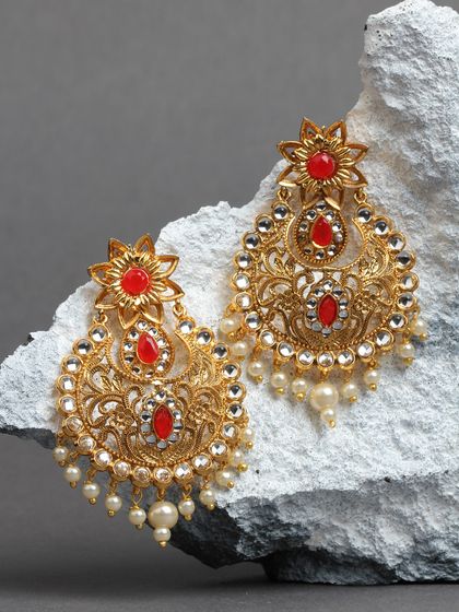 A pair of red and gold chandbalis resting on a stone. The intricate filigree work and vibrant red stones make these a perfect choice for a traditional event.