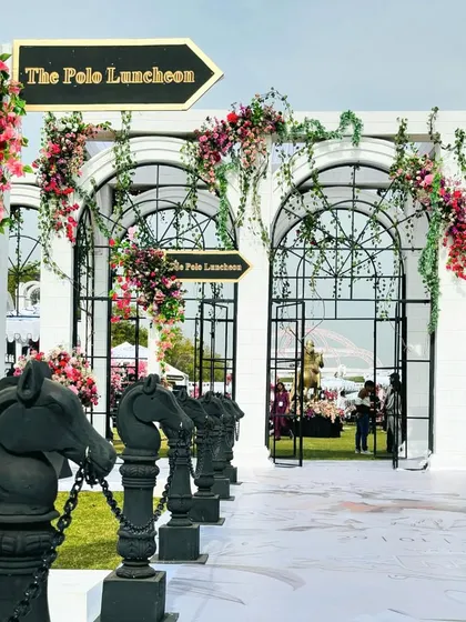 The grand entrance to 'The Polo Luncheon', inspired by 'My Fair Lady'. The walkway is lined with horse-head statues and flanked by white archways adorned with climbing pink and red flowers.