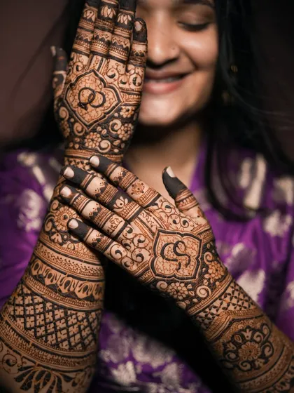A smiling client posing with her engagement mehndi, which features her initials and ring symbols.