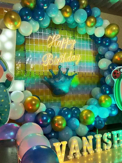 A first birthday celebration with a Cocomelon theme, using blue and green balloons, character cutouts, and a neon "Happy Birthday" sign.
