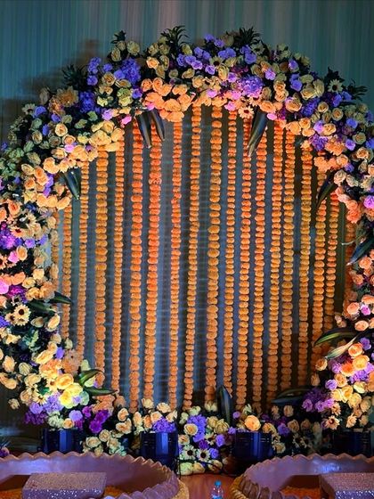A stunning Haldi backdrop featuring a large circular frame decorated with marigolds and purple flowers. The hanging marigold strings in the center create a traditional yet stylish look.