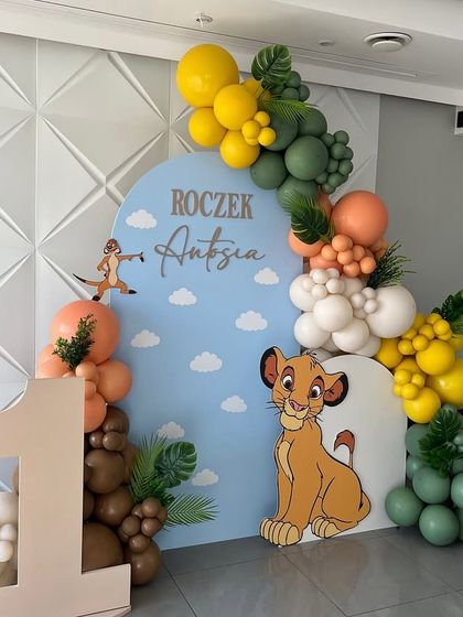A Lion King themed first birthday party. The setup features a custom backdrop with Simba, a balloon garland in jungle colors, and a large number '1' cutout.