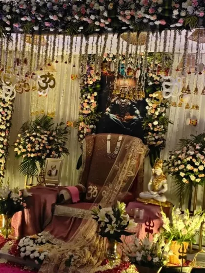 An elaborate gaddi setup for a darbar. The throne is decorated with rich fabrics and Om symbols, with a portrait of Lord Shiva in the background and flanked by large bouquets of fresh flowers.