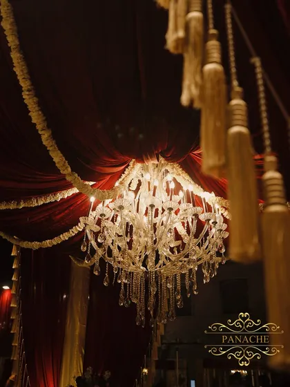 A close-up of a crystal chandelier against the rich maroon draped ceiling. The interplay of light and luxurious fabric is a signature element of our royal decor style.