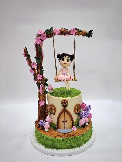 A fairytale cake featuring a girl on a swing under a blossoming tree, sitting atop a hobbit-house-style cake.