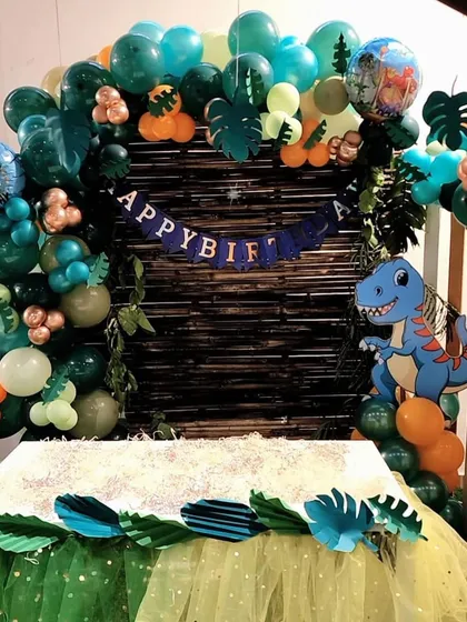 The cake table setup for a dinosaur party. The table skirt is decorated with paper leaves, and a "Happy Birthday" banner hangs above a rustic backdrop.