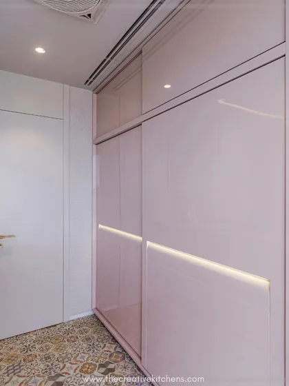 A full view of the glossy pink PU sliding wardrobe. The inbuilt shutter lights cast a mesmerizing glow, illuminating the narrow passage and making the space feel larger and more luxurious.