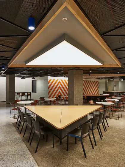 A unique triangular communal table in a cafeteria with an industrial aesthetic. The space features concrete pillars, exposed ceilings, and bold orange and white chevron patterns on the wall.