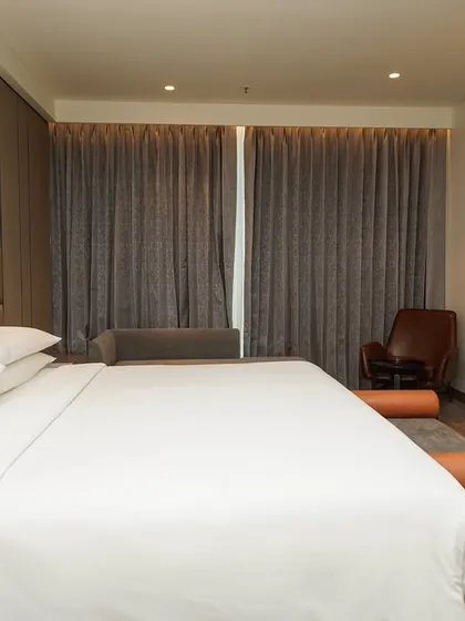 A close-up perspective of the neatly made bed in a modern hotel room, focusing on the crisp white linens and the textures of the headboard and bench.