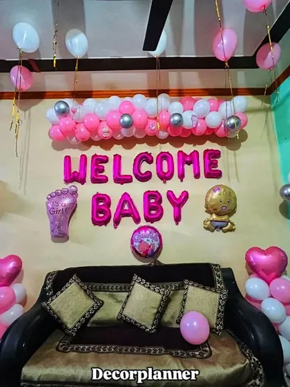 A full living room setup for a baby girl's welcome. The decor includes a balloon garland over the sofa, two balloon pillars, and foil letters, completely transforming the space for the special occasion.