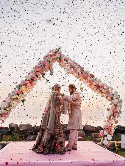 A wide shot of the Varmala ceremony by the sea, with confetti raining down. The open-air setting and the beautiful floral arch create a breathtaking scene for the couple's union.
