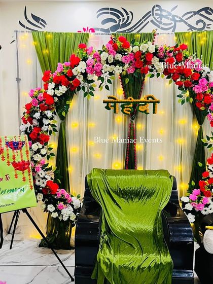 A vibrant Mehendi setup with a circular floral arch in red, pink, and white, set against a green and white draped backdrop.