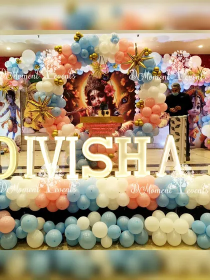 A grand stage for Divisha's Krishna-themed first birthday. The backdrop features an image of Little Krishna, framed by an elaborate balloon arch and large marquee letters.