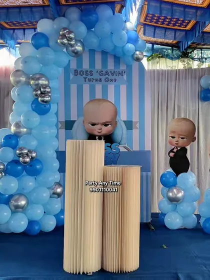 A Boss Baby theme for a first birthday. This setup includes character cutouts and a blue and silver balloon arch against a striped backdrop.