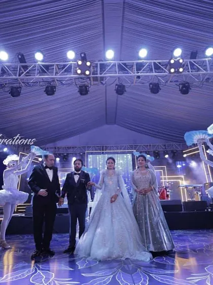 The couple stands center stage, framed by their troupe of whimsical dancers. This wide shot captures the full theatricality of the welcome I design for sangeet and reception nights.