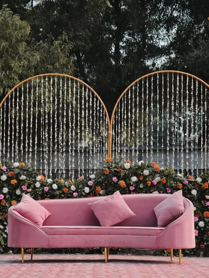 A beautiful floral stage setup with a pink sofa set against a backdrop of floral walls and hanging white garlands, creating a romantic and picturesque photo zone.