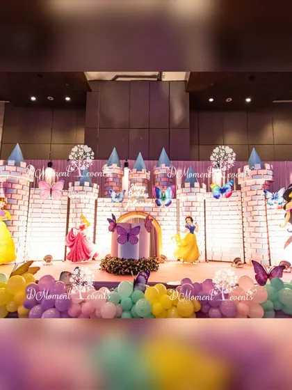 A fairytale castle stage setup for a princess-themed birthday party. The backdrop features a beautifully detailed castle wall with turrets, surrounded by character cutouts of Disney princesses like Snow White, Belle, and Jasmine.