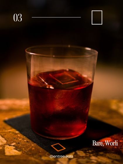 A classic dark cocktail, shot with warm, intimate lighting. The focus is on the rich color of the drink and the large, perfectly square ice cube, conveying a sense of quality and craftsmanship.