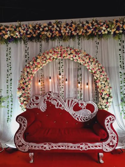 A classic stage decoration for an engagement or reception. The centerpiece is a beautiful floral arch over a royal red and silver sofa, creating a perfect spot for the couple.