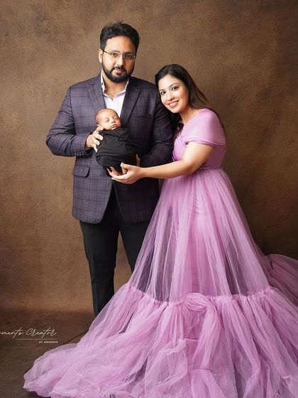 An elegant family portrait with their newborn son. The mother's flowing lavender gown and the classic brown backdrop create a sophisticated and timeless image.