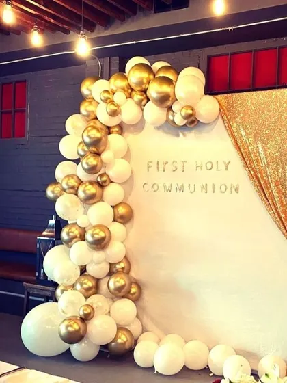 An elegant Holy Communion photo corner with a white backdrop and a gold sequin curtain. The scene is framed by a white and gold balloon garland.