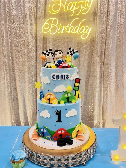 A grand two-tier car-themed cake for Chris's first birthday, set up at his party with a neon "Happy Birthday" sign.