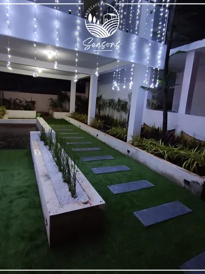 A modern residential landscape featuring an artificial lawn, stone slab pathway, and a raised planter bed. The string lights add a magical touch, making the space perfect for evening relaxation.