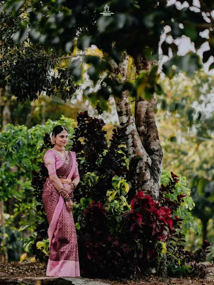 A graceful portrait of a bride in a pink saree, standing amidst lush greenery, showcasing her calm and elegant presence.
