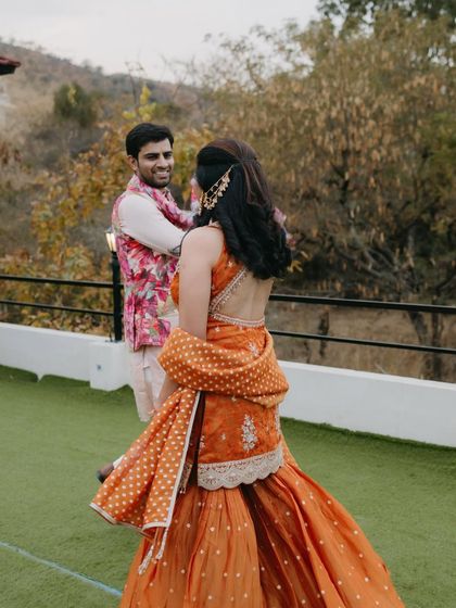 A lovely moment between the couple at their sundowner mehendi. This shot shows how his floral jacket kurta set complements her traditional orange sharara, creating a beautiful, coordinated picture.