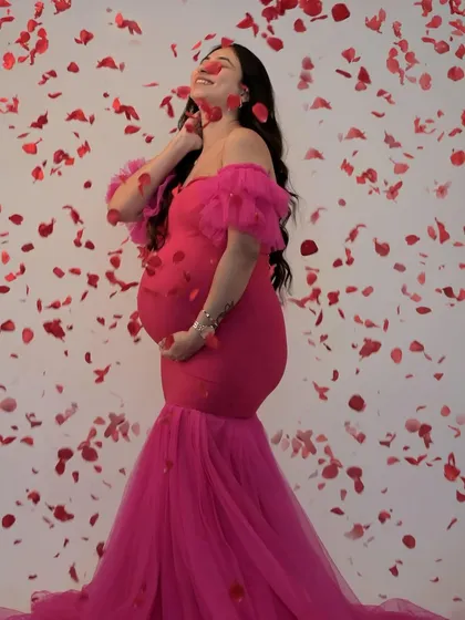 Celebrate your joy with this vibrant hot pink mermaid gown. The off-shoulder ruffle sleeves and form-fitting style beautifully highlight your baby bump, while the falling petals add a touch of playful romance to this studio shot.