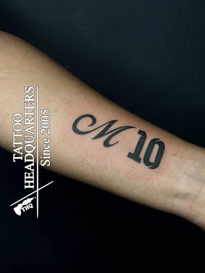 A custom "M 10" tattoo on the forearm, combining a stylized initial with a bold number for a personal and unique design.