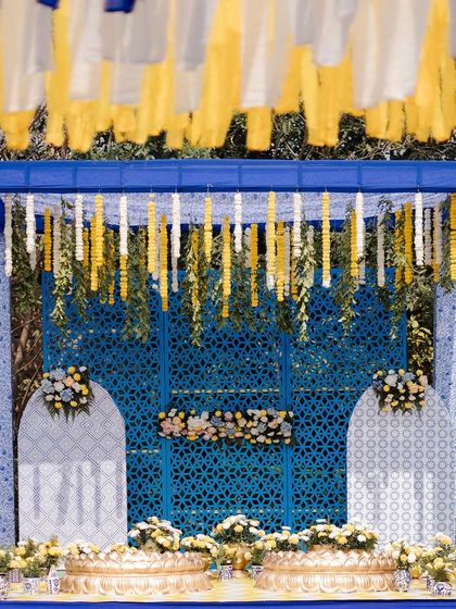 A striking Haldi stage in royal blue and yellow. The backdrop features intricate jaali panels and is framed by hanging floral strings, with traditional brass urlis for the ceremony.