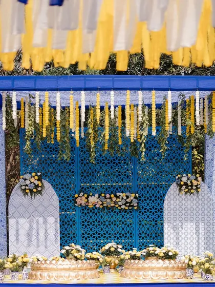 A striking Haldi stage in royal blue and yellow. The backdrop features intricate jaali panels and is framed by hanging floral strings, with traditional brass urlis for the ceremony.
