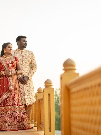 A candid moment of a couple in ornate traditional wedding attire, looking out from a palace balcony. The warm light adds a soft, romantic glow.