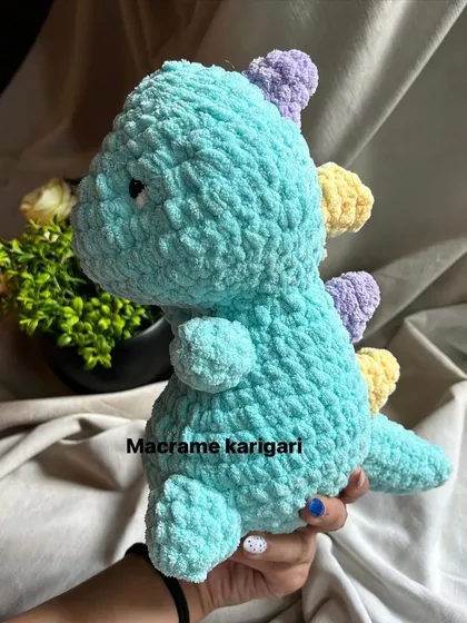 Roar! This adorable turquoise dinosaur is made from soft chunky yarn with pastel-colored spikes. It's a friendly dino perfect for any child's toy collection.