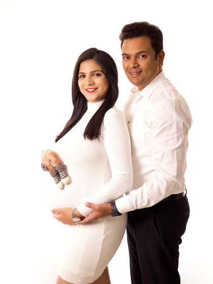 A classic high-key studio portrait. The couple's matching white outfits and the bright background create a timeless and joyful image, with the tiny baby shoes adding a sweet touch.