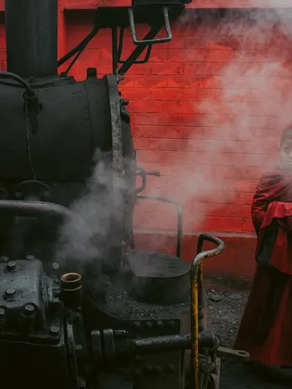 A wide, cinematic shot of a young monk in Darjeeling, standing beside a steam train. The 16x9 frame emphasizes the dramatic composition and colors.