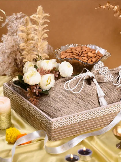 This rustic and elegant Diwali hamper features a woven tray, jute potlis, a bowl of almonds, and beautiful floral accents, creating a warm and sophisticated gift.