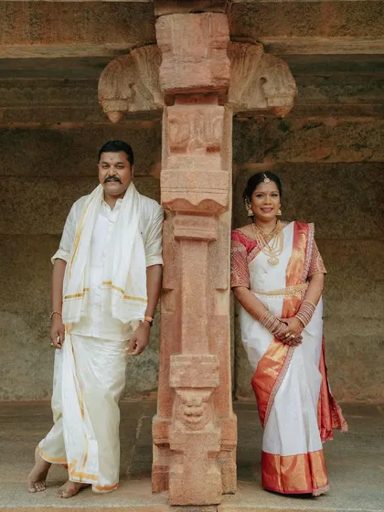 A classic portrait of a couple in traditional South Indian wedding attire. They are posed against a carved stone pillar, which adds a touch of heritage and cultural significance to the photo.