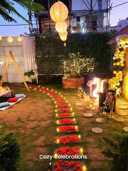 A beautiful garden party layout at night. A candle-lit path of red flowers leads through the lawn, connecting a boho picnic area to a balloon arch, all under a canopy of paper lanterns and string lights.
