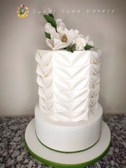 This two-tiered white wedding cake features a modern, geometric folded fondant technique on the top tier. It's complemented by a beautiful spray of handcrafted white sugar magnolias, creating a design that is both contemporary and timeless.