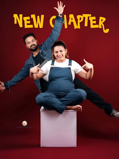 A high-energy and fun couple's portrait. The partner jumps excitedly behind the seated mother-to-be, both pointing to the bump under the banner 'New Chapter'.