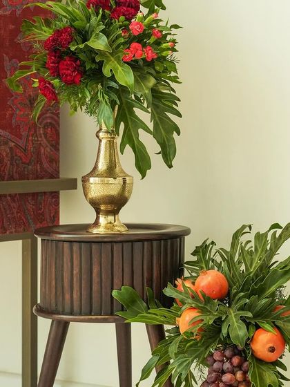 A corner detail from 'दाड़म ka Bageecha', with a brass vase holding red flowers and a lower arrangement of pomegranates and grapes, set against a rich, patterned fabric.
