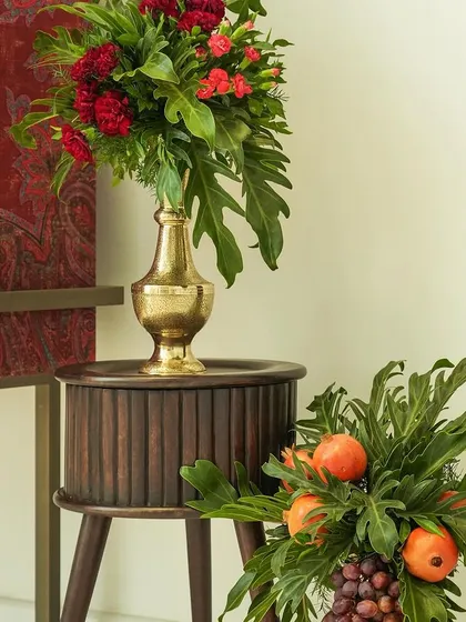 An elegant corner arrangement from the event. A tall brass vase with red flowers sits atop a wooden side table, while a lower arrangement of pomegranates and greenery provides balance.